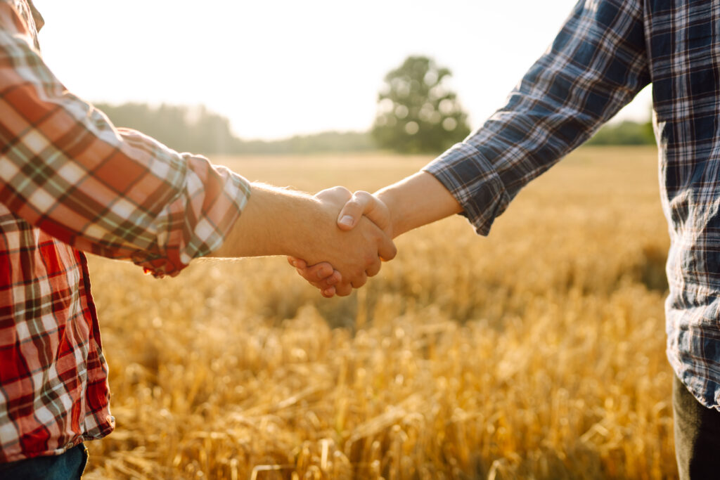 Dois agricultores apertam as mãos no contexto de um campo de trigo ao pôr do sol.