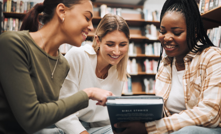 Três mulheres leem um livro em uma biblioteca